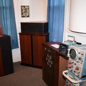 Vintage loudspeakers on wooden cabinets on display in museum room with white walls and blue curtains