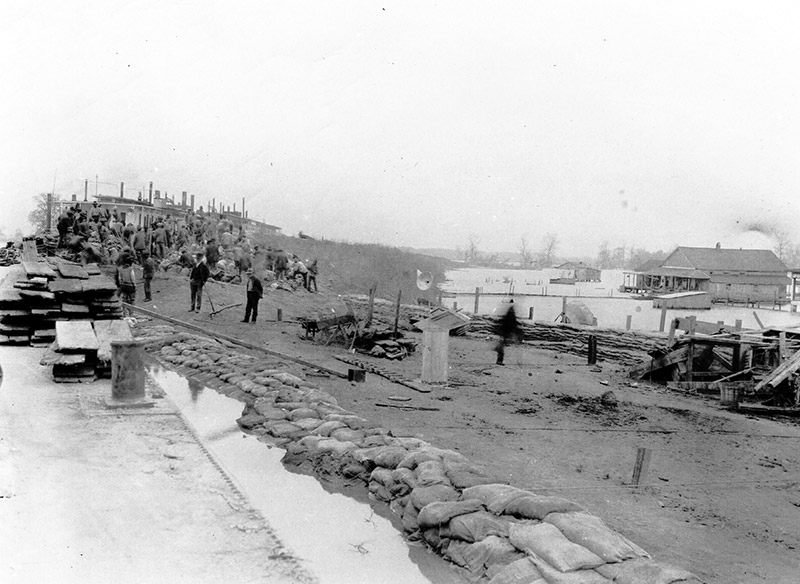 Group of men and stacks of sandbags on flooded shore with farm under water nearby
