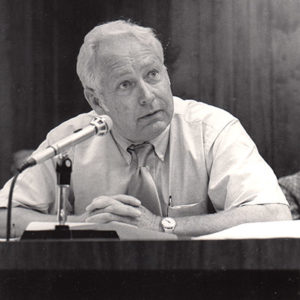 White man sitting in shirt and tie speaking into a microphone