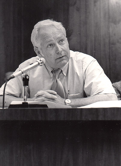 White man sitting in shirt and tie speaking into a microphone