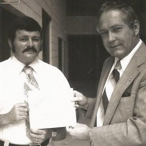 White man in suit and tie handing award to white man with mustache in shirt and tie