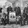 Group of white men in suits with portrait of white man in suit
