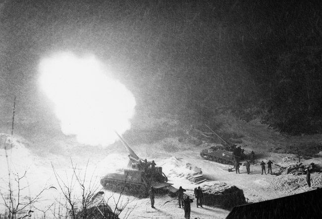 Two artillery cannons, one of them firing into the air, with soldiers covering their ears amid heavy snow