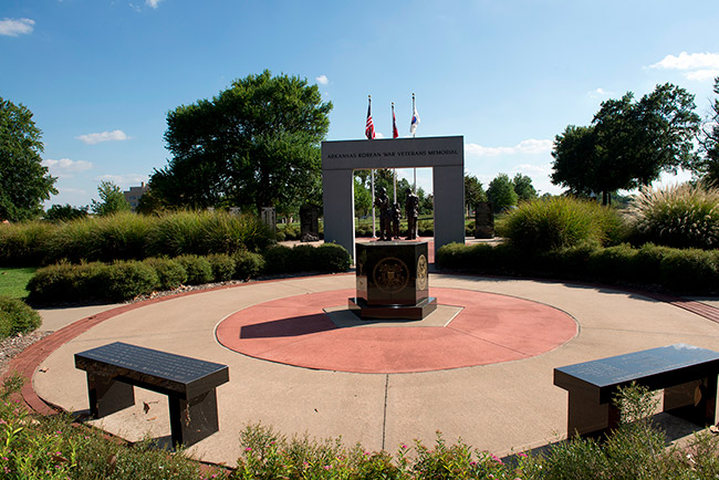 Round concrete platform with two benches and statue in center and concrete entrance way with three flag poles