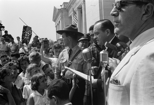 White man in suit speaking into microphones outside classical stone building with guard and crowd with american flag and confederate flag