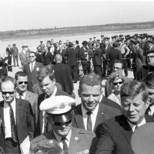 White man in suit and tie talking to a man in military uniform with people around them and spectators looking on behind a rope