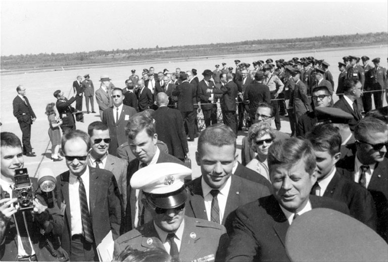 White man in suit and tie talking to a man in military uniform with people around them and spectators looking on behind a rope