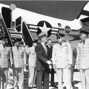 Mixture of white men in military uniforms and other white men and one woman in civilian suits standing at base of airplane