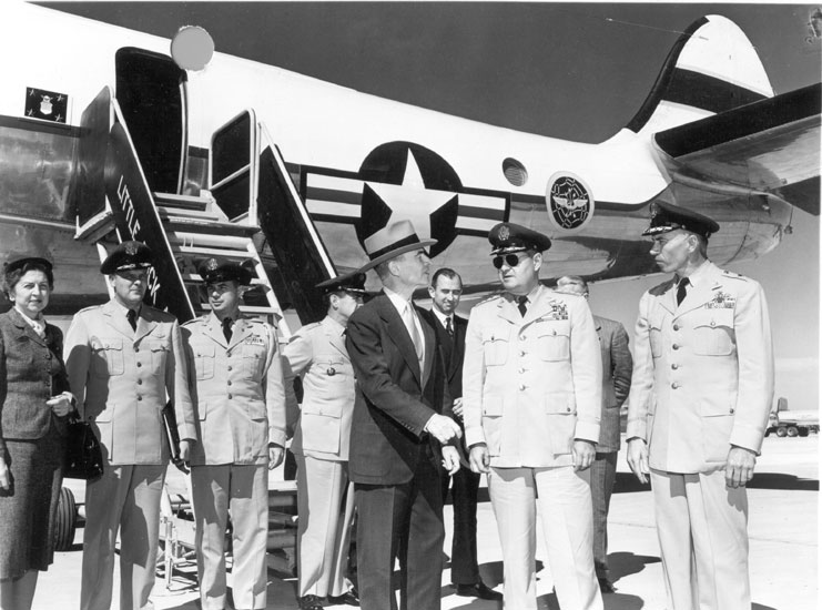 Mixture of white men in military uniforms and other white men and one woman in civilian suits standing at base of airplane