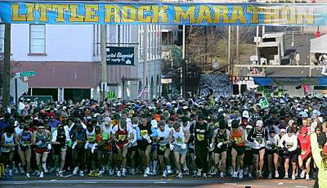 Mixed crowd of people in shorts standing on city street with "Little Rock Marathon" banner hanging above them