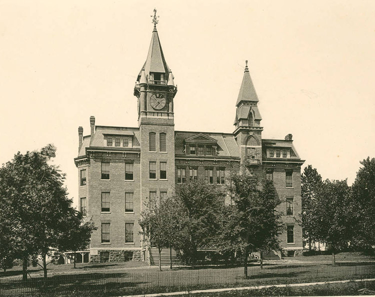 Multistory brick building with clock tower and second tower with trees in its front yard