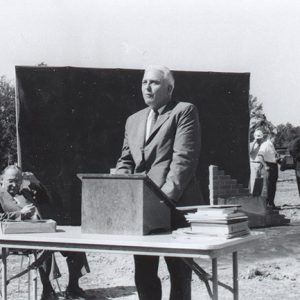 White man in suit speaking at table with lectern on it with white man sitting beside him and white men standing behind him