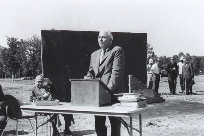 White man in suit speaking at table with lectern on it with white man sitting beside him and white men standing behind him