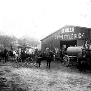 Men and horse-drawn carriages in front of barn with "Stables City of Little Rock" sign