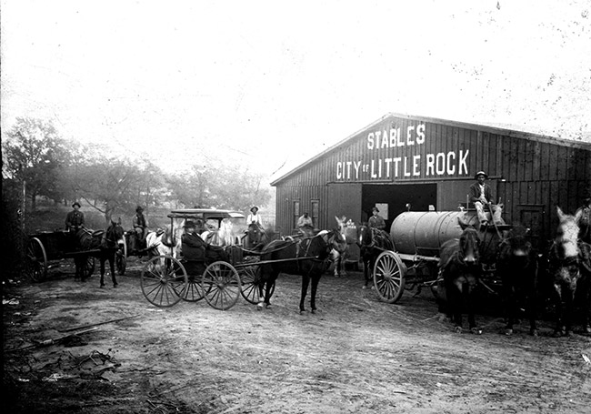 Men and horse-drawn carriages in front of barn with "Stables City of Little Rock" sign