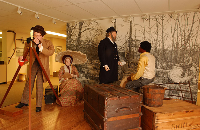 Museum display with mannequins in period costume as a white surveyor, woman with parasol, steamboat captain, and black man