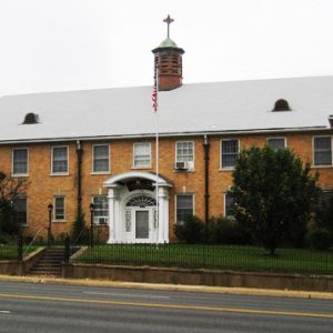 Two-lane road next to multistory building with cupola and cross on its roof inside iron fence with trees