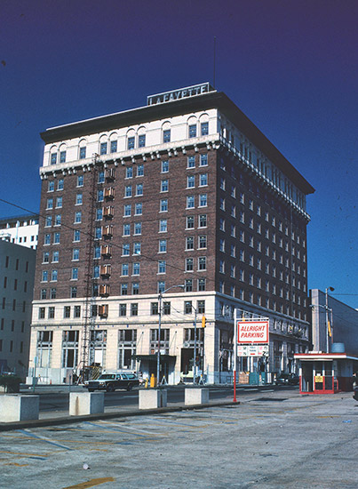 Tall hotel building on street corner with "LaFayette" sign on top next to a paid parking lot with red trimmed booth