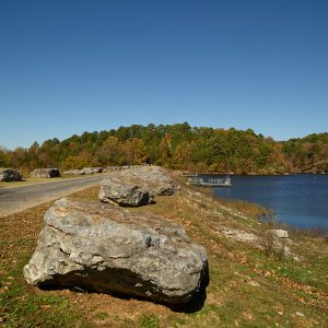 Paved walking path lined with rocks next to lake with fishing docks on it and tree covered hills in the background