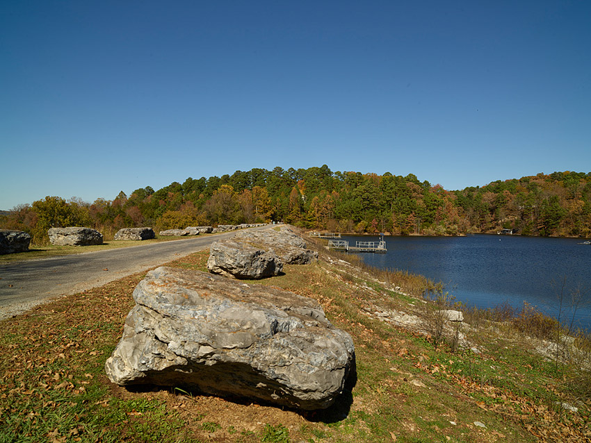 Paved walking path lined with rocks next to lake with fishing docks on it and tree covered hills in the background