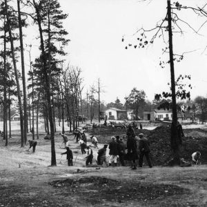 Group of men digging in field with single-story buildings and houses in the background