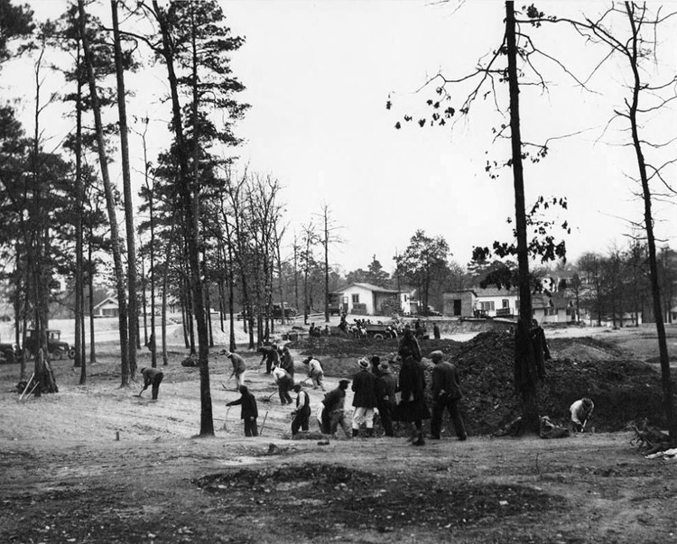 Group of men digging in field with single-story buildings and houses in the background