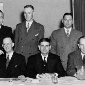 Five white men in suit and tie sitting at a table with three white men standing behind them