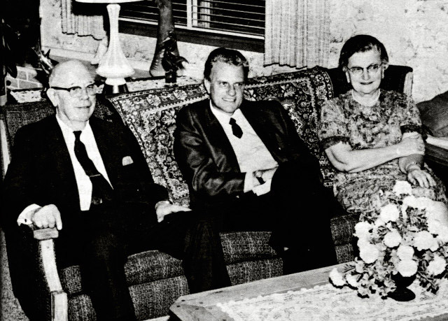 Older white men in suits sitting on a couch with white woman