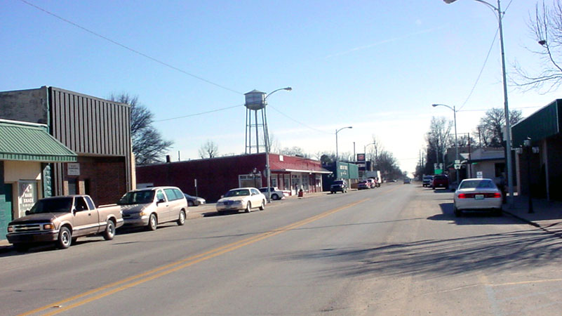 Town street lined with buildings and parked cars with trees and water tower in the background