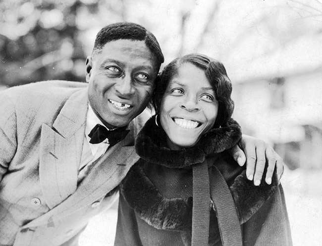 African-American man in suit who has a gap in his teeth with his arm around a smiling woman wearing a fur-trimmed coat