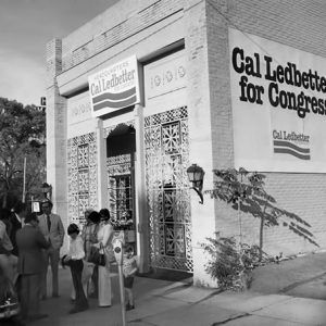 Group of people standing outside brick storefront with lattice details with "Cal Ledbetter for Congress" signs above the door and on side wall