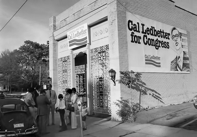 Group of people standing outside brick storefront with lattice details with "Cal Ledbetter for Congress" signs above the door and on side wall