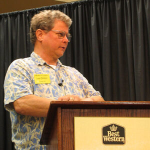 White man with glasses in Hawaiian shirt speaking at lectern