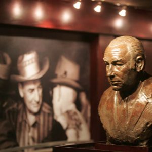Bust of man displayed in room with wood paneled walls and spotlighted photograph of white men in hats