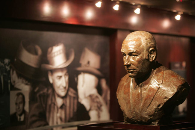 Bust of man displayed in room with wood paneled walls and spotlighted photograph of white men in hats