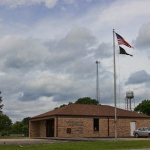 Brick post office building on parking lot and flag pole with radio tower and water tower in the background