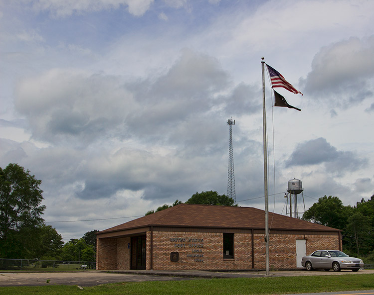 Brick post office building on parking lot and flag pole with radio tower and water tower in the background