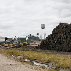 Stack of lumber and industrial buildings on gravel with water tower in the background