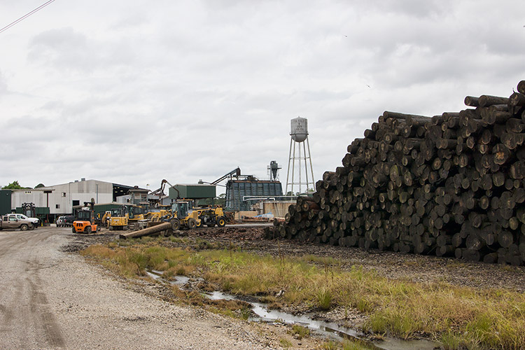Stack of lumber and industrial buildings on gravel with water tower in the background
