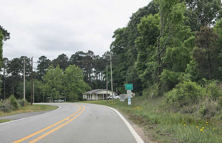 Green "Leola" sign on right side of two-lane road with single-story houses in the background