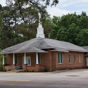Brick church building with covered entrance and steeple on two-lane street