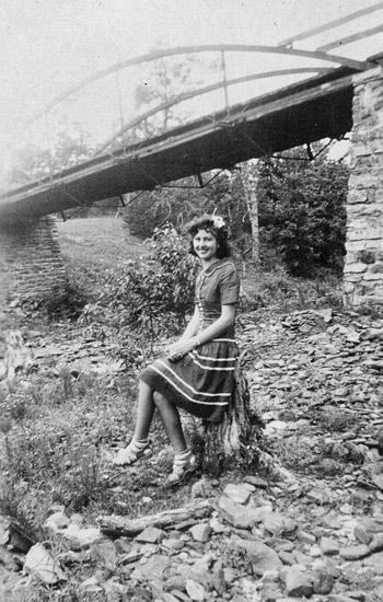 White woman with curly hair in striped dress sitting on a tree stump with bridge behind her