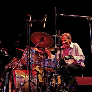 Older white man in white shirt and black pants smiling behind drum set on stage