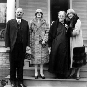 White man in a suit and tie with two white women in fur coats and old woman in a black dress
