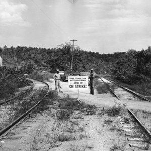 Two male workers standing between two sets of railroad tracks with picket sign