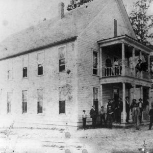 White men women and children outside two-story hotel with covered porch and balcony