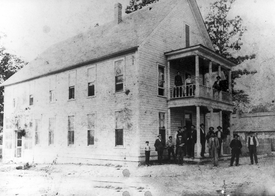 White men women and children outside two-story hotel with covered porch and balcony