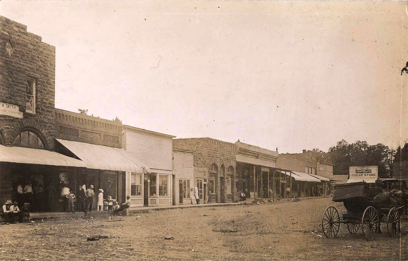 People on sidewalk outside single and multistory buildings on dirt street with horse drawn carriage in the foreground