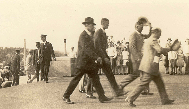 Group of white men walking with police and crowd watching in the background