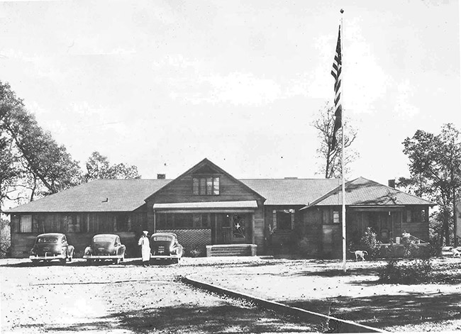 Faded black and white lodge building with three cars and flag pole in front yard and person standing by one car
